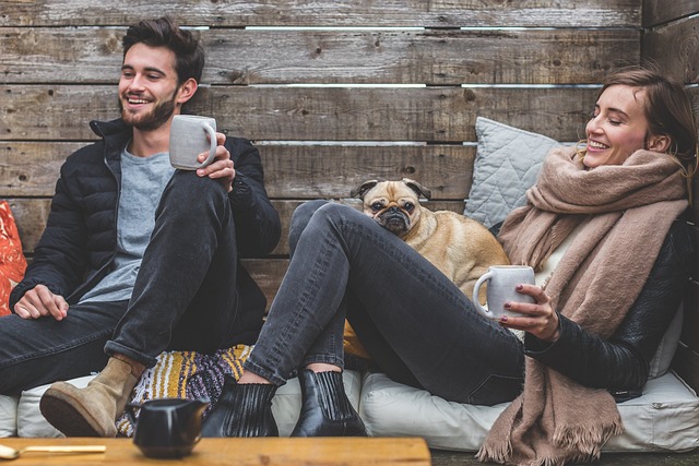 Auf dem Foto befinden sich ein Mann und eine Frau in einer einfachen Holzhütte. Beide halten eine Tasse in der Hand, sie lächeln. Die Frau hat einen kleinen Hund auf ihrem Schoß.